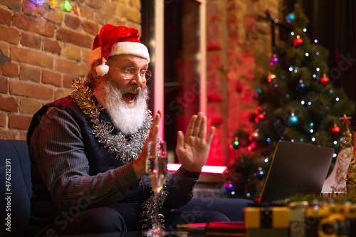 Emotional old bearded man sitting at home and celebrates New Year with friends and family by means of video call.