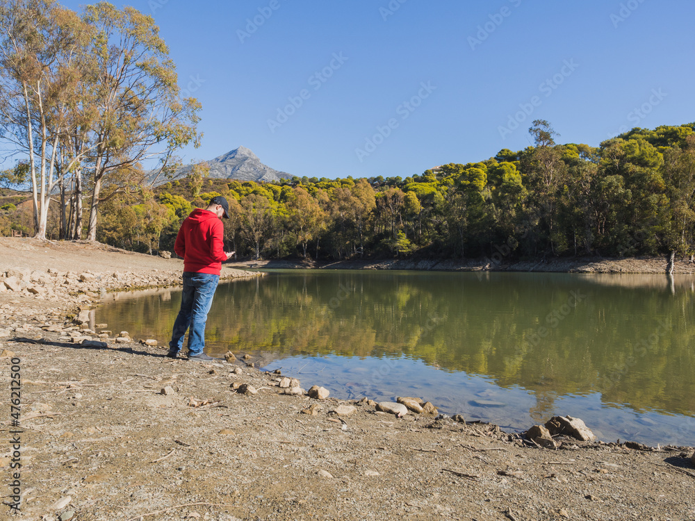 A young man in a red sweatshirt looks at smartphone next to a lake and with natural landscape in the background