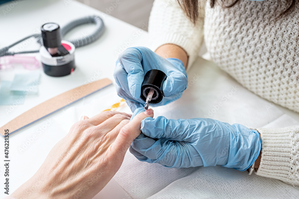 Manicure master in mask and gloves putting on gel polish on the nails of a client