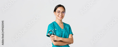 Covid-19, coronavirus disease, healthcare workers concept. Professional good-looking asian doctor, medical worker in glasses and scrubs, cross arms and smiling, white background
