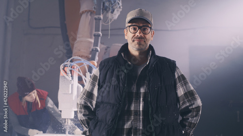 Self assured mature male artisan in casual outfit with glasses and cap holding hands on waist and looking at camera while standing near woman cutting polymer blocks with robotic arm milling machine