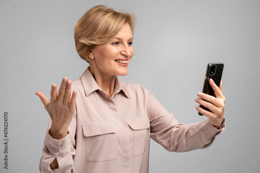 Portrait of cheerful woman in casual shirt taking selfie using smartphone, talking on video call and smiling joyfully, mobile communication. Indoor studio shot, grey background