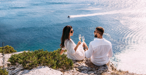 A couple in love drinking champagne on the seashore, panorama. A happy couple on a honeymoon by the sea, panorama. A beautiful couple with champagne on the coast. Honeymoon. Wedding travel. Copy space © MISHA