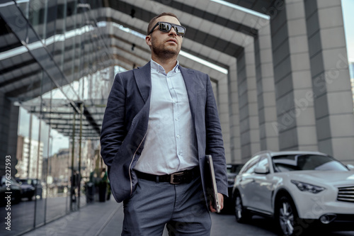 Young businessman in sunglasses holding his laptop. Walking on the street. © Vlad