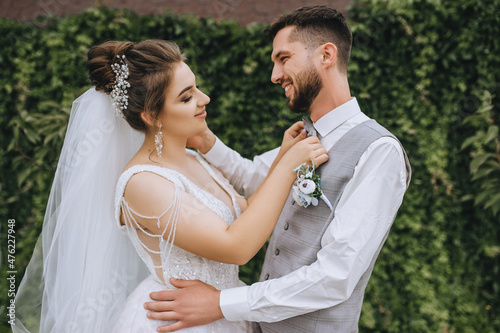 A beautiful, sweet bride in a white dress straightens a bow tie to her beloved groom in a gray suit against the background of blooming green decorative grapes in nature. Wedding photography. photo