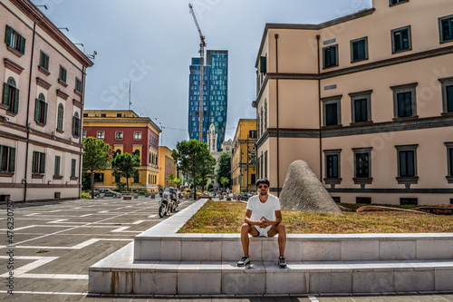 Young male tourist sits against the background of the Forever Green Tower skyscraper in Tirana, Albania. Beautiful street cityscape in the Albanian capital