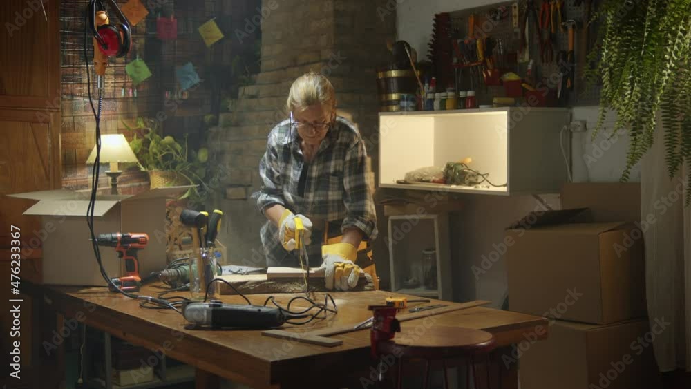 Mature woman carpenter cutting wood board with a saw in a small workshop, hair collected Mature woman carpenter cutting wood board with a saw in a small workshop, hair collected
