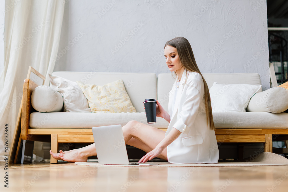 Beautiful young woman working on laptop computer while sitting at the living room, drinking coffee Beautiful young woman working on laptop computer while sitting at the living room, drinking coffee