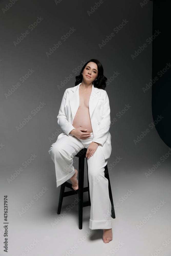 Caucasian young beautiful pregnant woman with dark hair in a white fashionable elegant pantsuit posing on a white background. isolates the banner space for text. Fashion for pregnant women. A business Caucasian young beautiful pregnant woman with dark hair in a white fashionable elegant pantsuit posing on a white background. isolates the banner space for text. Fashion for pregnant women. A business