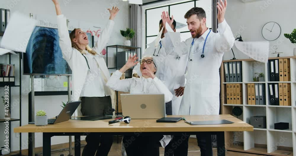 Attractive cheerful satisfied successful skilled multiethnic doctors recieving good news on laptop screen,they scattering reports over their heads,clapping hands and giving high five each other Attractive cheerful satisfied successful skilled multiethnic doctors recieving good news on laptop screen,they scattering reports over their heads,clapping hands and giving high five each other