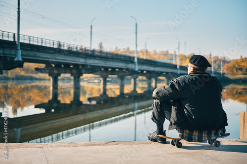 Back view of skater male relax sitting on skateboard at empty embankment on the urban background