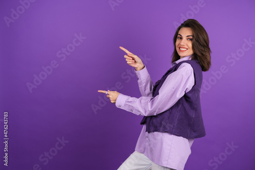 Young woman in a shirt and a vest on a purple background smiles cheerfully in high spirits, points a finger at an empty space to the left