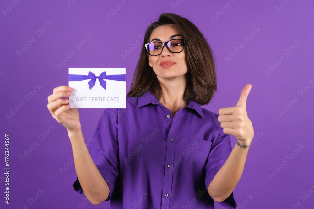 Young woman in a shirt on a purple background with a gift certificate smiling cheerfully, with makeup, lipstick on her lips and glasses, excited