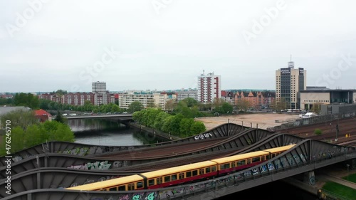 Fly over steel bridge over river in city. Suburban train running on multitrack railway line. Residential neighbourhood in background. Berlin, Germany