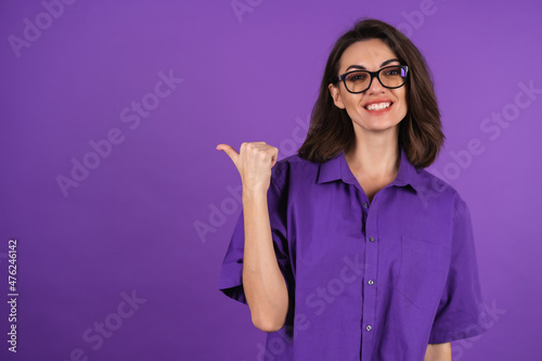 Young woman in a shirt with short sleeves on a purple background, with makeup and stylish glasses, points a finger to the left with a smile