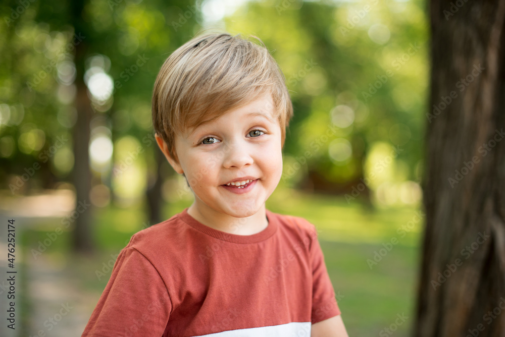Joyful little boy. He is smiling on the background of a green park. The boy looks into the camera lens Joyful little boy. He is smiling on the background of a green park. The boy looks into the camera lens
