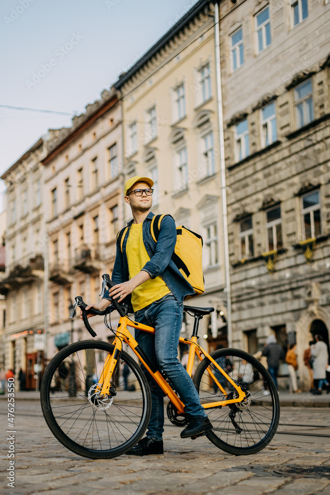 A happy delivery man who is sitting on a bicycle and looking around. He is carrying a yellow backpack on his shoulders. Vertical photo.