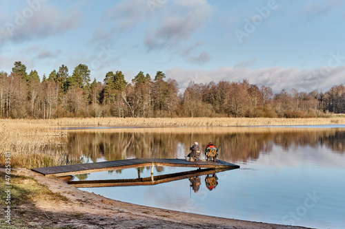 Man and woman with fishing rods sitting on wooden bridge among scenic lake, reflecting on mirror surface of water.