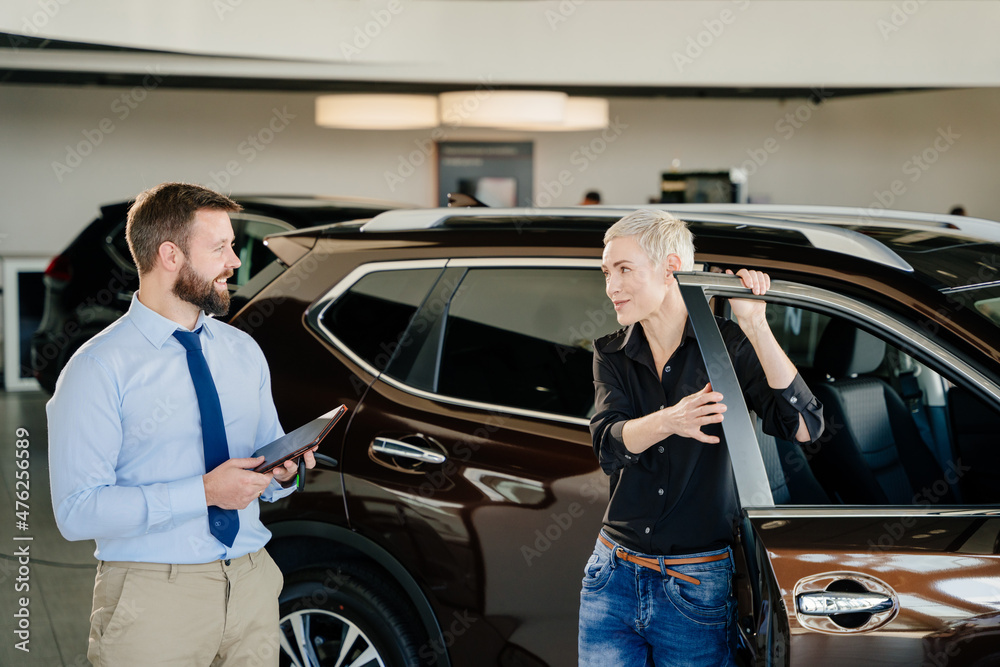 Adult female customer and smiling male car dealer standing at car salon interior. Smiling friendly man car seller with tablet in hands talking to a woman customer in black shirt who wants to buy car.