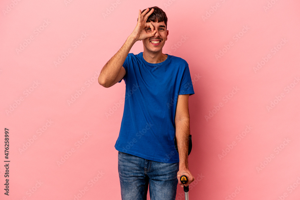 Young mixed race man with crutches isolated on pink background excited keeping ok gesture on eye. Young mixed race man with crutches isolated on pink background excited keeping ok gesture on eye.