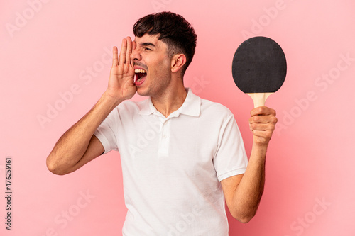 Young mixed race man holding a ping pong racket isolated on pink background shouting and holding palm near opened mouth.
