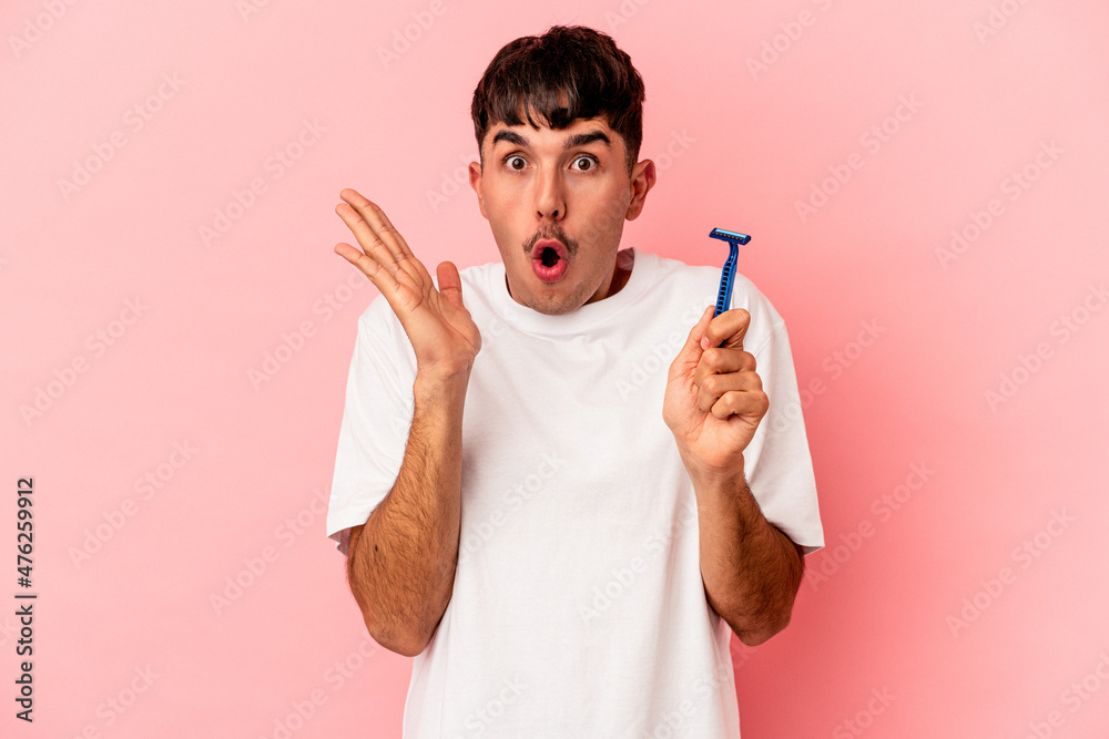 Young mixed race man holding razor blade isolated on pink background surprised and shocked.