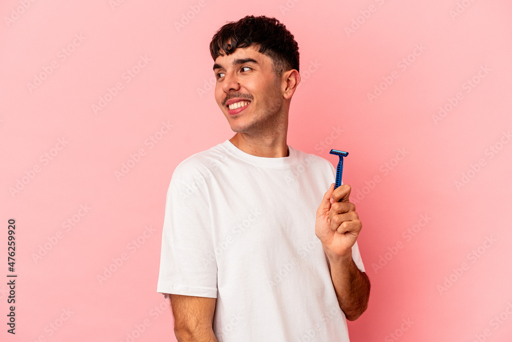 Young mixed race man holding razor blade isolated on pink background looks aside smiling, cheerful and pleasant. Young mixed race man holding razor blade isolated on pink background looks aside smiling, cheerful and pleasant.