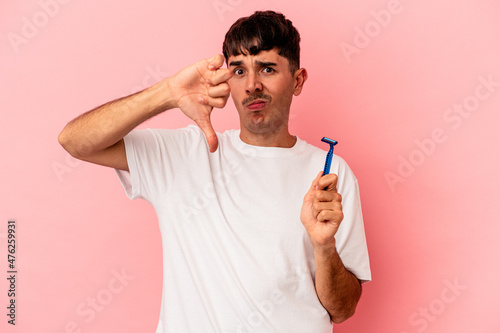 Young mixed race man holding razor blade isolated on pink background showing a dislike gesture, thumbs down. Disagreement concept.