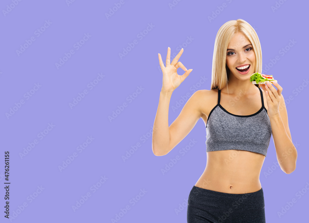 Woman in sportswear holding сrispbread sandwich with cheese, tomato and salad, showing okay, isolated over violet purple background. Slim blond girl at studio shot. Health, beauty and dieting concept. Woman in sportswear holding сrispbread sandwich with cheese, tomato and salad, showing okay, isolated over violet purple background. Slim blond girl at studio shot. Health, beauty and dieting concept.