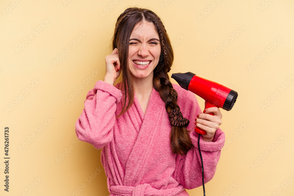 Young woman holding a hairdryer isolated on yellow background covering ears with hands. Young woman holding a hairdryer isolated on yellow background covering ears with hands.