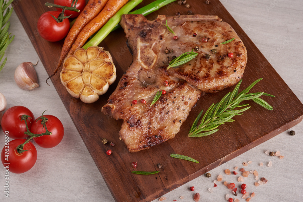 Grilled pork steak from a summer BBQ served with Vegetables, asparagus, baby carrots, fresh tomatoes and spices. Grilled steak on wooden cutting board on stone background . top view. Grilled pork steak from a summer BBQ served with Vegetables, asparagus, baby carrots, fresh tomatoes and spices. Grilled steak on wooden cutting board on stone background . top view.