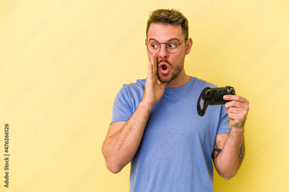 Young caucasian man holding a game controller isolated on yellow background is saying a secret hot braking news and looking aside Young caucasian man holding a game controller isolated on yellow background is saying a secret hot braking news and looking aside