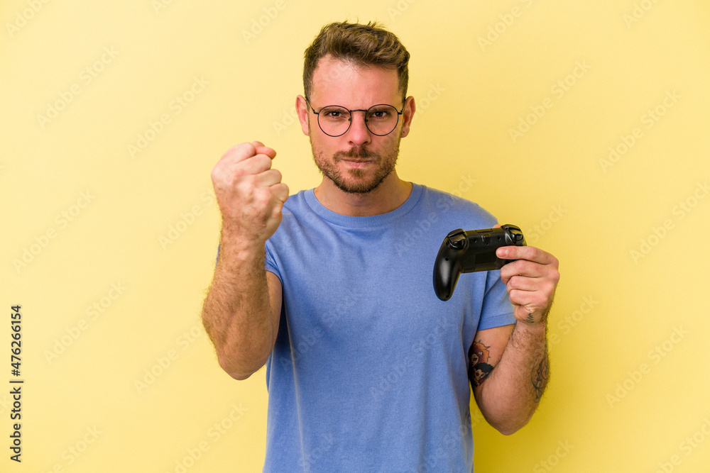 Young caucasian man holding a game controller isolated on yellow background showing fist to camera, aggressive facial expression. Young caucasian man holding a game controller isolated on yellow background showing fist to camera, aggressive facial expression.