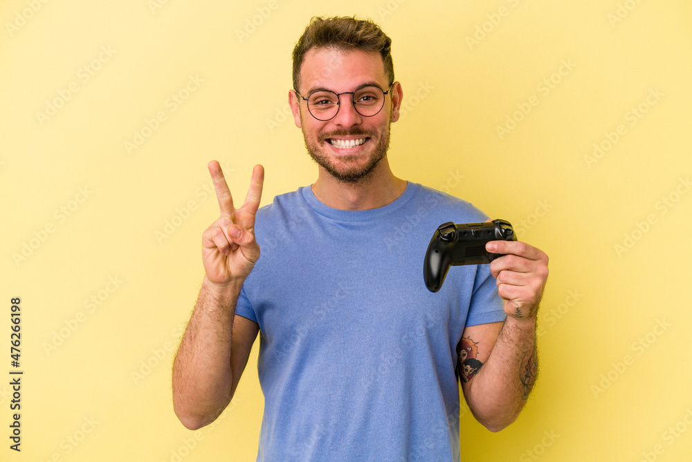Young caucasian man holding a game controller isolated on yellow background showing number two with fingers. Young caucasian man holding a game controller isolated on yellow background showing number two with fingers.