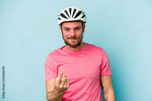 Young caucasian man with bike helmet isolated on blue background pointing with finger at you as if inviting come closer.