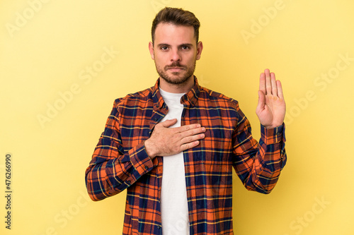 Young caucasian man isolated on yellow background taking an oath, putting hand on chest.