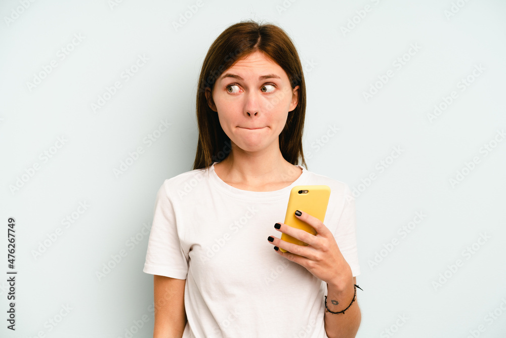 Young English woman holding mobile phone isolated on blue background confused, feels doubtful and unsure. Young English woman holding mobile phone isolated on blue background confused, feels doubtful and unsure.