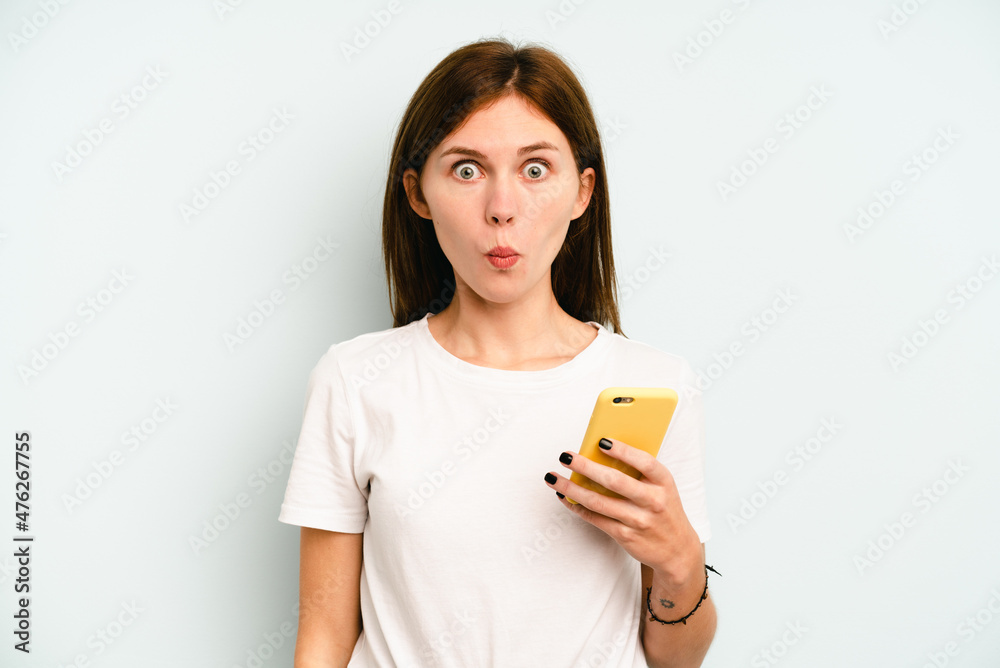 Young English woman holding mobile phone isolated on blue background shrugs shoulders and open eyes confused. Young English woman holding mobile phone isolated on blue background shrugs shoulders and open eyes confused.
