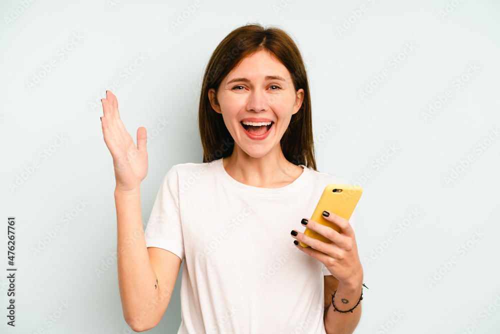 Young English woman holding mobile phone isolated on blue background receiving a pleasant surprise, excited and raising hands. Young English woman holding mobile phone isolated on blue background receiving a pleasant surprise, excited and raising hands.