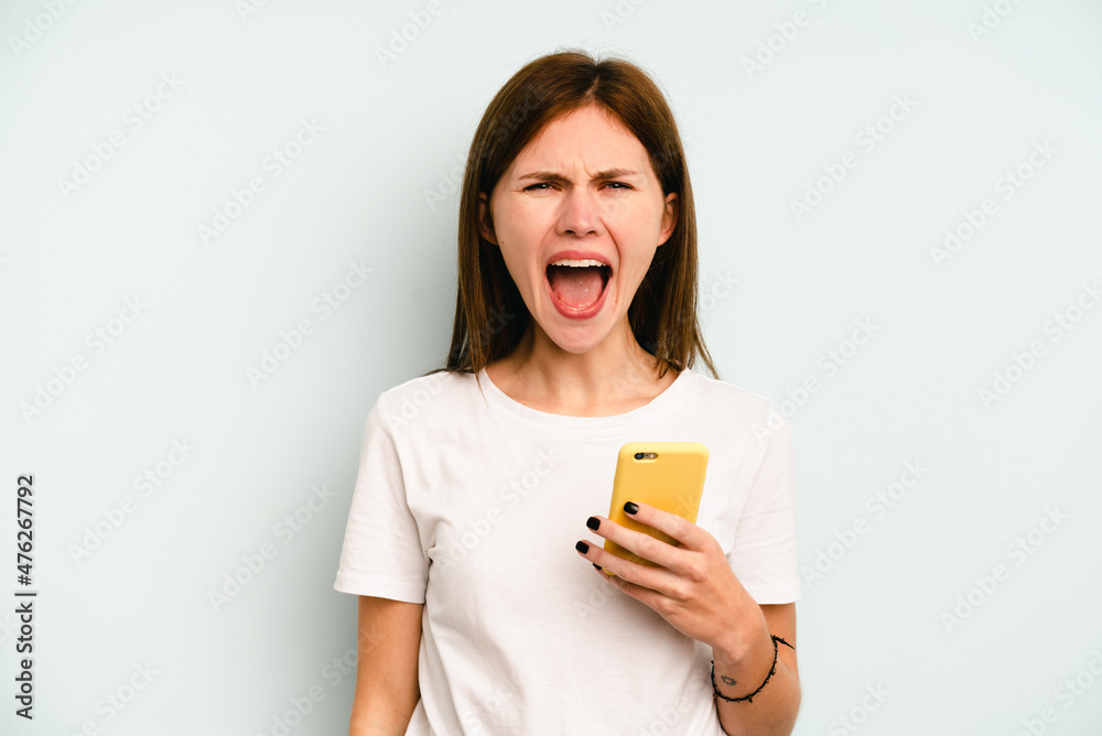 Young English woman holding mobile phone isolated on blue background screaming very angry and aggressive. Young English woman holding mobile phone isolated on blue background screaming very angry and aggressive.
