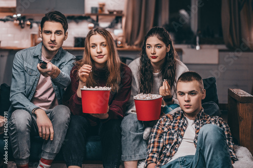 A company of cute young two guys and two girls sitting on the couch, watching TV, eating popcorn, bored, changing the channel