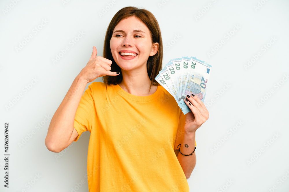 Young English woman holding banknotes isolated on blue background showing a mobile phone call gesture with fingers. Young English woman holding banknotes isolated on blue background showing a mobile phone call gesture with fingers.
