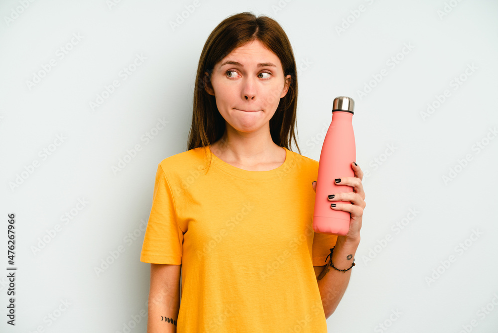 Young English woman holding a thermo isolated on blue background confused, feels doubtful and unsure. Young English woman holding a thermo isolated on blue background confused, feels doubtful and unsure.