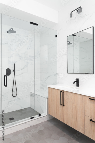 A beautiful luxury, modern bathroom with a light wood cabinet, walk-in shower with marble tiled walls, and black faucets and hardware.