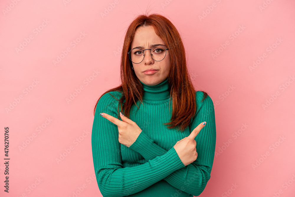 Fototapeta premium Young caucasian woman isolated on pink background points sideways, is trying to choose between two options.