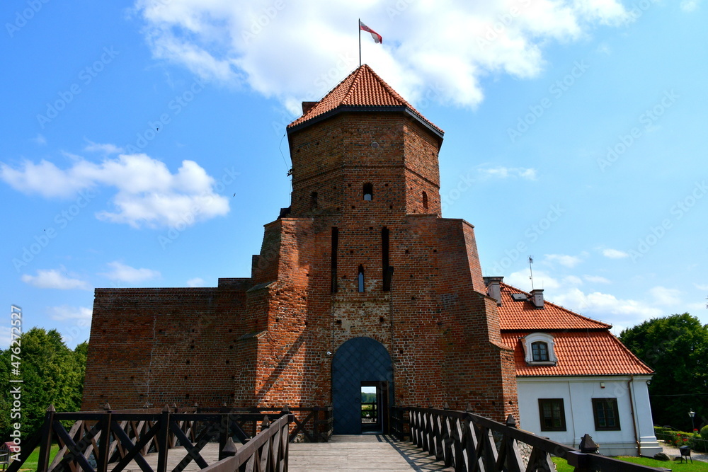 A view of an old medieval abandoned castle made out of red brick and with a Polish flag on top of its tower seen from the edge of a well made wooden bridge made out of planks, logs, and boards