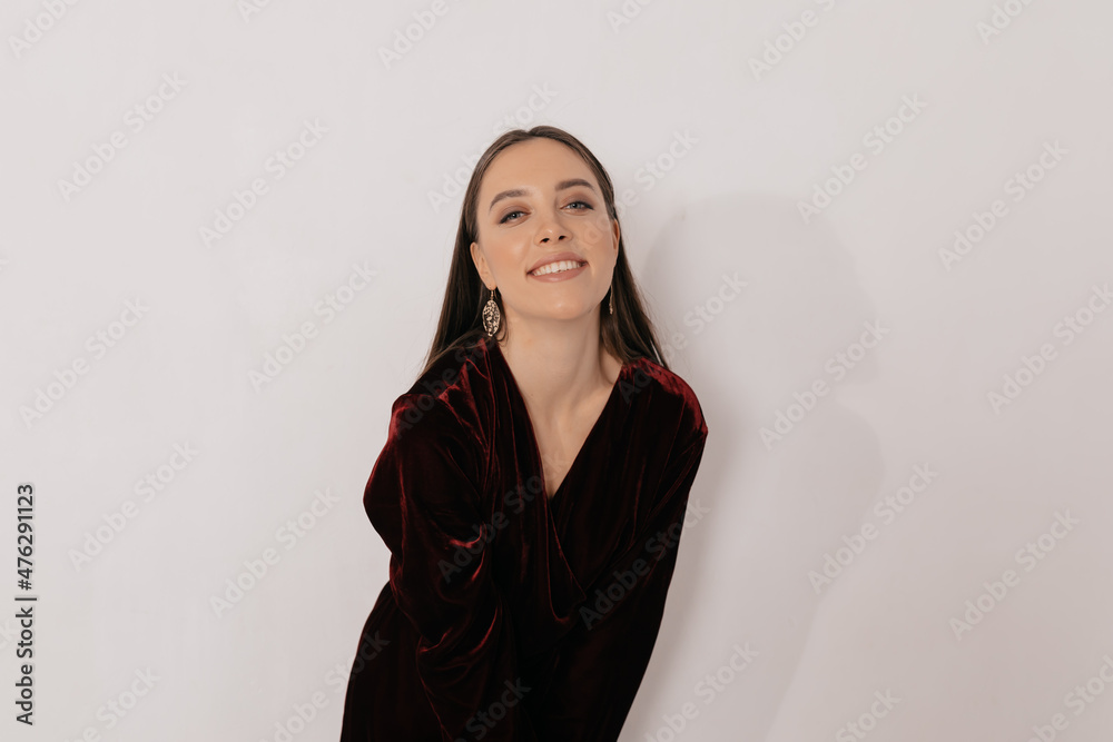 Fototapeta premium Charming adorable woman in stylish dress and wearing golden earnings smiling at camera over isolated backdrop. Calm happy brunette woman is looking at camera and laughing 
