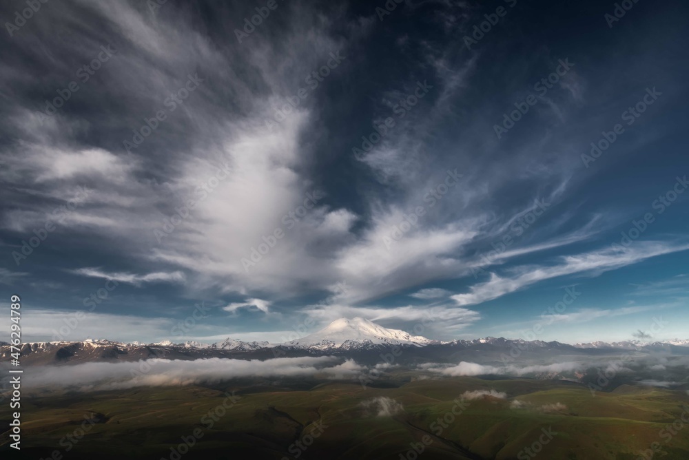 Stunning views of the highest two-peak mountain - Elbrus. There are epic white clouds in the blue sky. North Caucasus. View from the Kanzhol plateau. Stunning views of the highest two-peak mountain - Elbrus. There are epic white clouds in the blue sky. North Caucasus. View from the Kanzhol plateau.