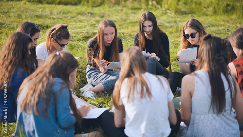 A group of female students are sitting in a circle on a meadow for collective work with notebooks.