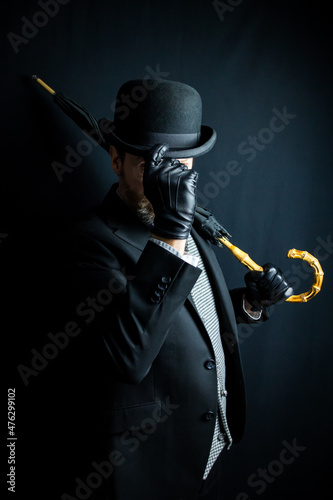 Portrait of Man in Dark Suit Holding Umbrella Over His Shoulder and Doffing Bowler Hat on Black Background. Classic English Gentleman.